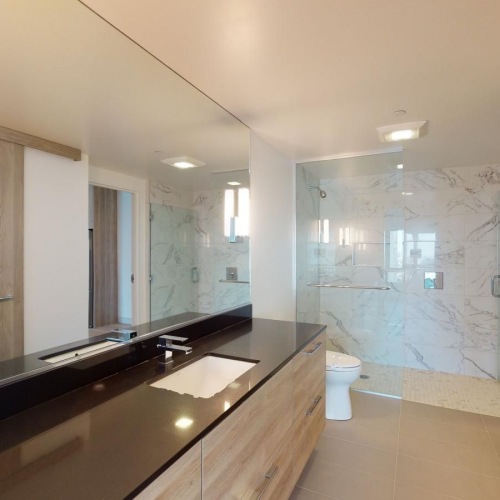 San Francisco, CA Luxury Apartments - Trinity Place at 33 8th - Beautiful Bathroom with a Glass Shower, Marble WAlls, a Large Vanity with Black Countertops, Wooden Cabinetry, and a Large Mirror